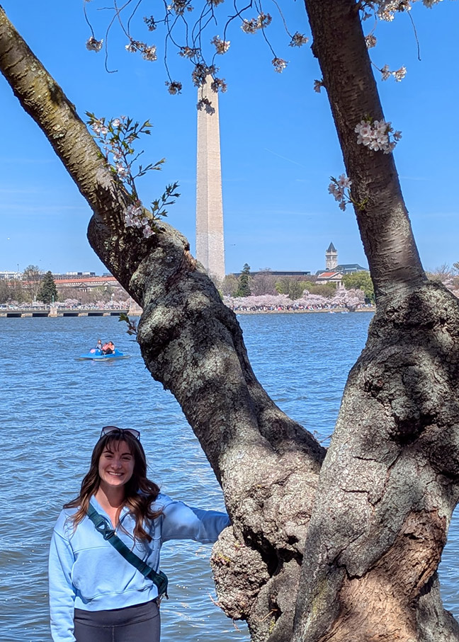 Sierra Bradtz standing next to an old cheery blossom tree with the reflection pool and Washington Monument behind her