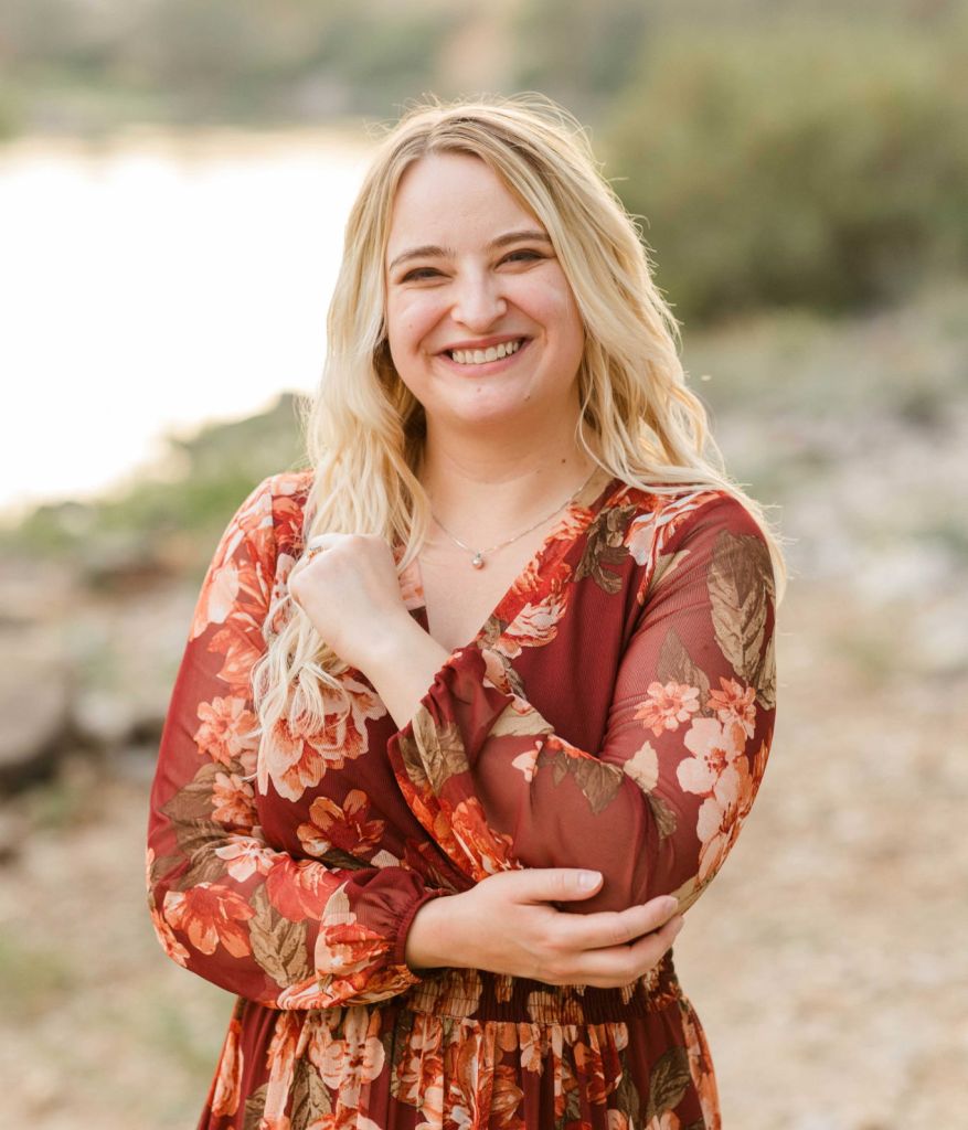 Sandra Spearman. Young woman with chest length blonde hair, standing near water and smiling at the camera. She is wearing a flowy, maroon floral dress.