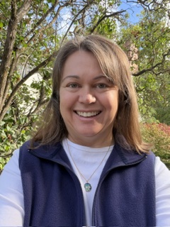 Kristie Lawrence. White woman with shoulder length brown hair, wearing a white shirt and blue vest.
